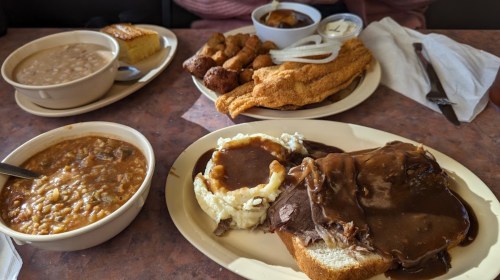 A table with a variety of comfort foods: roast beef with gravy, mashed potatoes, cornbread, and fried fish.