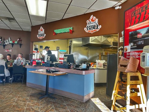 Interior of a BBQ restaurant with a counter, kitchen area, and customers seated at tables. Decor includes BBQ-themed signs.