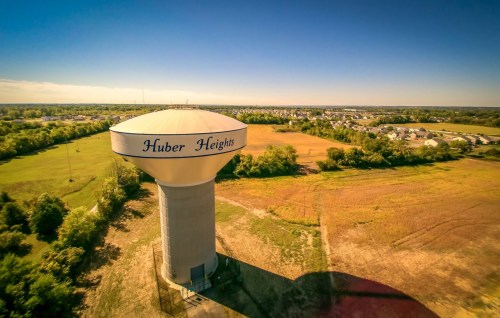 Aerial view of a water tower labeled "Huber Heights" surrounded by fields and a suburban area under a clear blue sky.