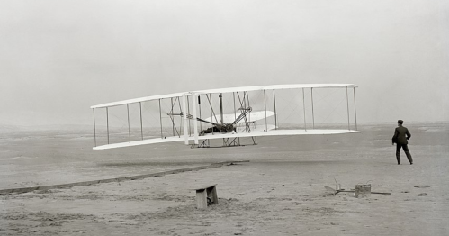 A historic biplane takes off from a sandy area, with a man observing in the background.