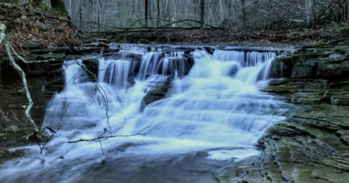 A serene waterfall cascades over rocky terrain, surrounded by trees and a tranquil forest setting.