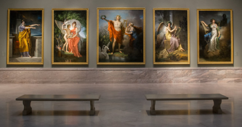 Five large, framed paintings displayed in a gallery, with two wooden benches in front on a polished floor.