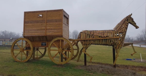 A wooden sculpture of a horse pulling a wagon, made from stacked wooden planks, set in a grassy area.