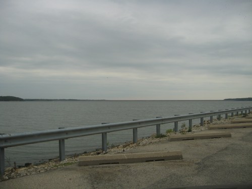 A calm lake under a cloudy sky, with a guardrail and parking area in the foreground.