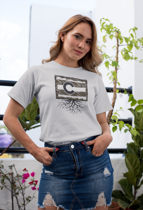 A woman in a gray t-shirt with a graphic design stands in front of plants, wearing a denim skirt.