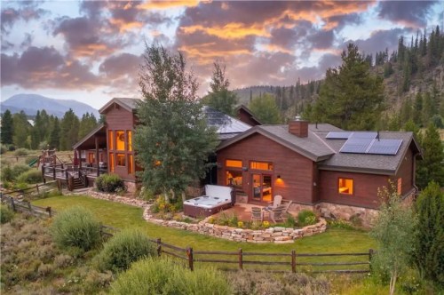 A modern wooden house with solar panels, surrounded by greenery and mountains, under a colorful sunset sky.