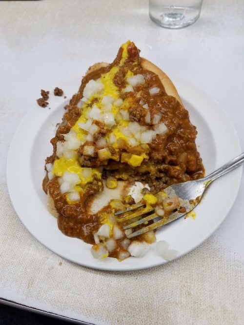 A plate with a half-eaten hot dog topped with chili, mustard, and diced onions, alongside a fork.