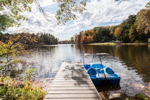 A serene lake scene with a wooden dock and a blue paddle boat, surrounded by autumn foliage and a cloudy sky.