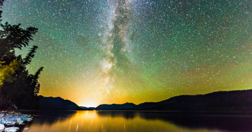 A serene lake reflects a starry sky with the Milky Way, surrounded by mountains and trees under a glowing horizon.