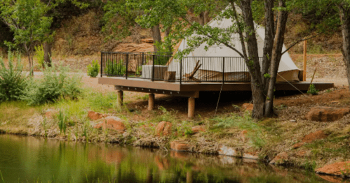 A glamping tent on a wooden deck by a serene pond, surrounded by trees and rocky terrain.