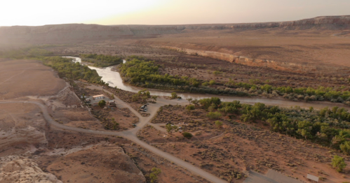 Aerial view of a winding river surrounded by green trees in a desert landscape with rocky cliffs in the background.