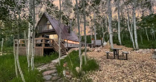 A cozy A-frame cabin surrounded by aspen trees, with a picnic area and a path leading to the entrance.
