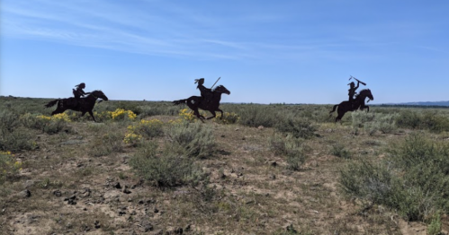 Silhouettes of three horsemen riding through a grassy landscape under a clear blue sky.