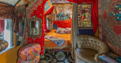 A cozy, colorful room with ornate textiles, a bed draped in vibrant fabrics, and a plush chair.