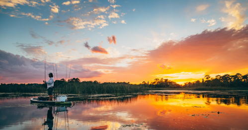 A person fishing on a calm lake at sunset, with vibrant colors reflecting on the water and lush greenery in the background.