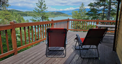 Two red lounge chairs on a wooden deck overlooking a serene lake surrounded by trees and mountains.