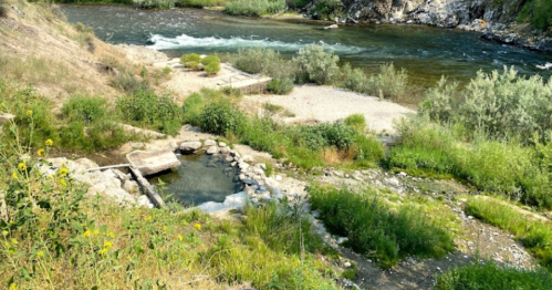 A serene riverside scene featuring a natural hot spring surrounded by lush greenery and flowing water.