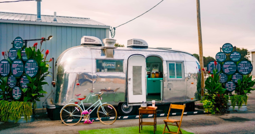 A shiny silver Airstream trailer with an "OPEN" sign, surrounded by plants and a bicycle, set in an outdoor space.