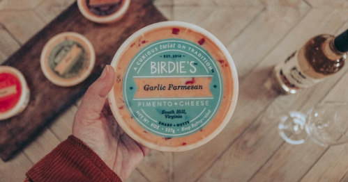 A hand holding a container of Birdie's Garlic Parmesan cheese, with a wooden board and wine bottle in the background.