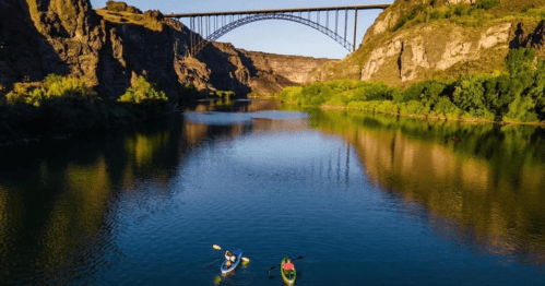 Two kayakers paddle on a calm river beneath a large bridge, surrounded by rocky cliffs and lush greenery.