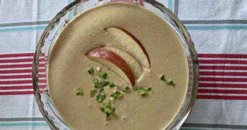 A bowl of creamy soup topped with apple slices and chopped green onions, set on a striped tablecloth.