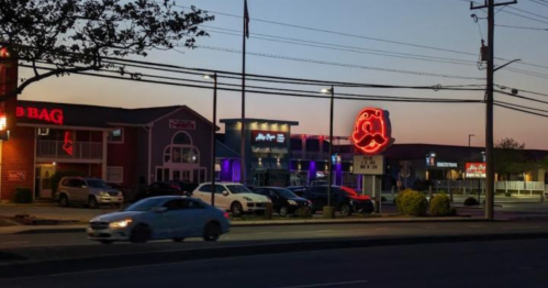A street view at dusk featuring neon signs, parked cars, and a mix of restaurants and shops.