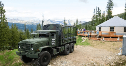 An army truck parked near a yurt, with mountains in the background and people enjoying outdoor activities.