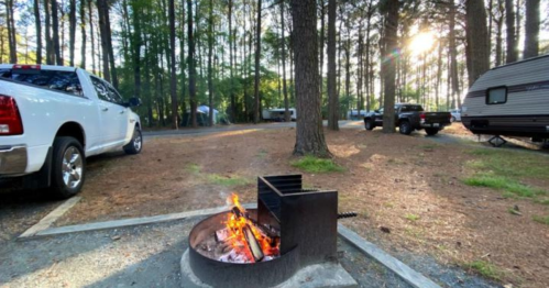A campfire burns in a fire pit surrounded by trees, with parked vehicles and campers in the background.