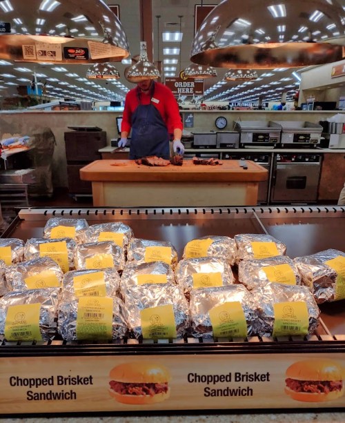 A man in an apron prepares chopped brisket sandwiches at a deli counter, with wrapped sandwiches displayed in front.