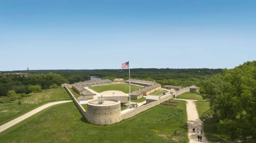 Aerial view of a historic fort with an American flag, surrounded by green fields and a clear blue sky.