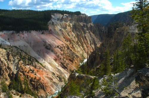 A scenic view of a colorful canyon with steep cliffs, lush trees, and a winding river below under a blue sky.