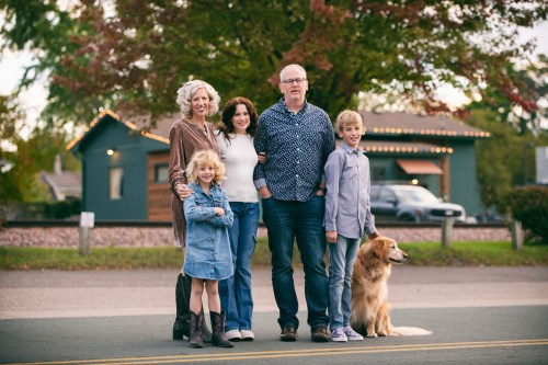 A family of five stands together on a street, smiling, with a golden retriever sitting beside them.