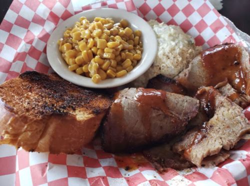 A plate of barbecue meat with sauce, a side of corn, creamy coleslaw, and toasted bread on a red-checkered paper.