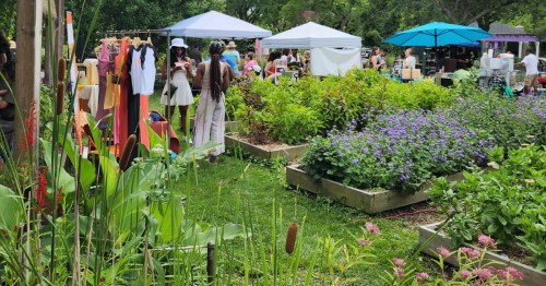 People browsing outdoor garden stalls and raised flower beds under white and blue umbrellas.