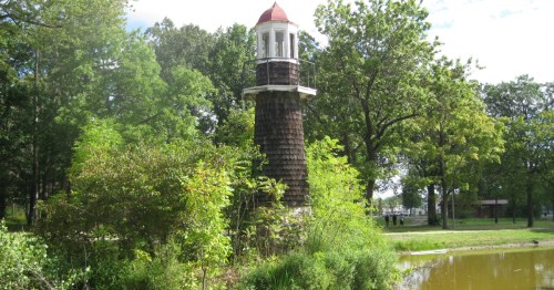 A rustic, shingle-covered decorative lighthouse surrounded by green bushes and trees near a pond.