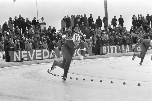 A speed skater races on an ice track, with a cheering crowd in the background, in a black and white photo.