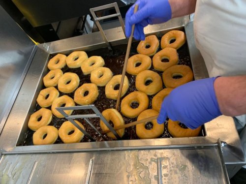 A person in gloves is frying donuts in a large fryer, with several golden donuts floating in hot oil.