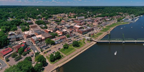 Aerial view of a riverside town with green parks, buildings, and a marina along the water's edge.