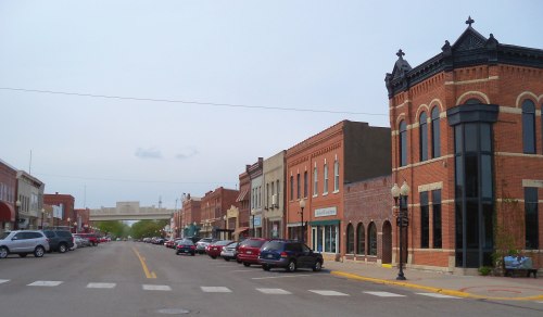 A view of a small town street lined with brick buildings and parked cars under a cloudy sky.