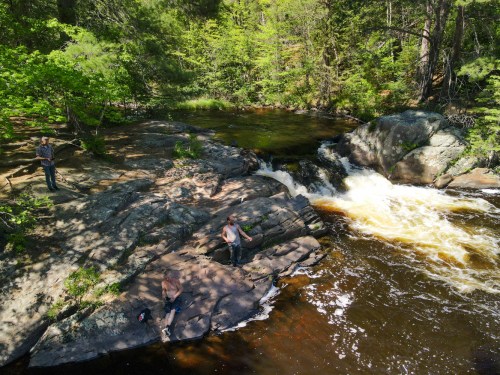 Two people relax on rocks by a river, while another stands nearby, surrounded by lush greenery and flowing water.