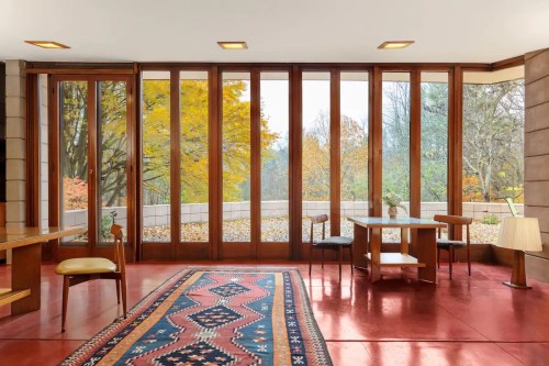 Interior of a modern room with large windows, wooden furniture, and a colorful rug, showcasing autumn foliage outside.