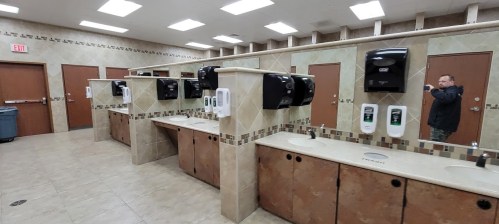 A clean public restroom with multiple sinks, soap dispensers, and hand dryers, featuring a person in the background.