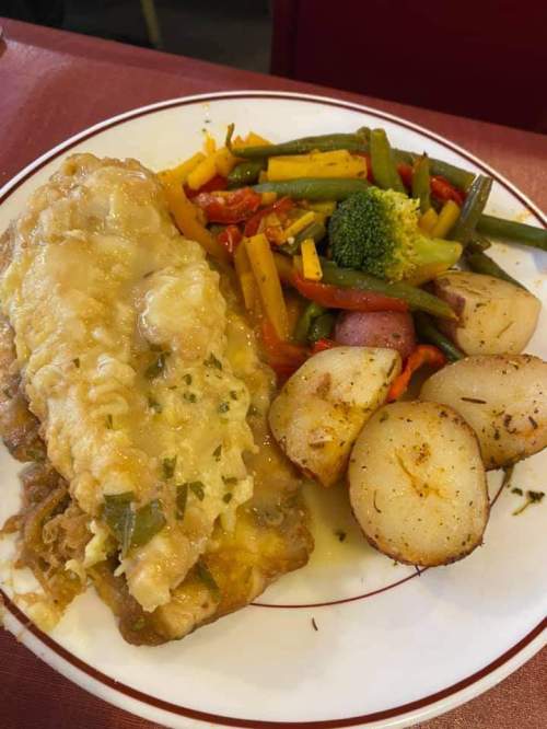 A plate of baked fish topped with cheese, served with mixed vegetables and roasted potatoes.