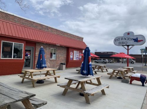 Outdoor seating area at Fat Racks BBQ, featuring picnic tables and umbrellas, with a sign and cloudy sky in the background.
