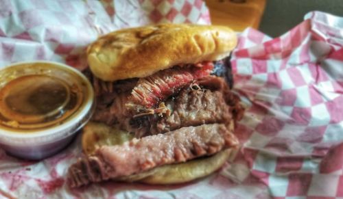 A close-up of a sandwich filled with sliced beef, served with a small cup of sauce, all on checkered paper.