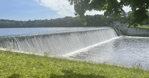 A serene view of a dam with water cascading over the edge, surrounded by greenery and a calm lake.