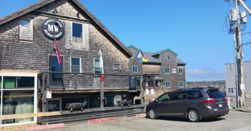 A wooden building with flags, a parked minivan, and a clear blue sky in a coastal setting.