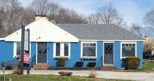 A blue building with a white roof, featuring an "Open" sign and a red "For Sale" banner in front.