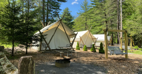 Two glamping tents surrounded by trees, with a picnic table, fire pit, and swings in a serene outdoor setting.