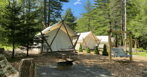 Two glamping tents surrounded by trees, with a picnic table, fire pit, and swings in a serene outdoor setting.